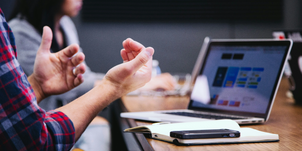 A software consultant sits at a conference table with their laptop open, arms gesturing as if explaining. They also have a notebook and a smartphone. The image is cropped to just show the arms. In the background, others sit at laptops.