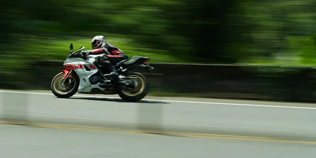 A person riding a white and red sport motorcycle races up a road with greenery in the background. The photo is blurry, as the camera and the rider are both moving.