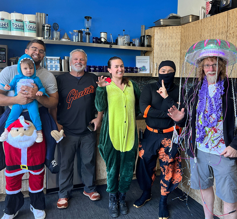 Digital foundry software engineers, consultants and staff pose in the breakroom in halloween costumes, one holding his baby who is dressed as a shark.