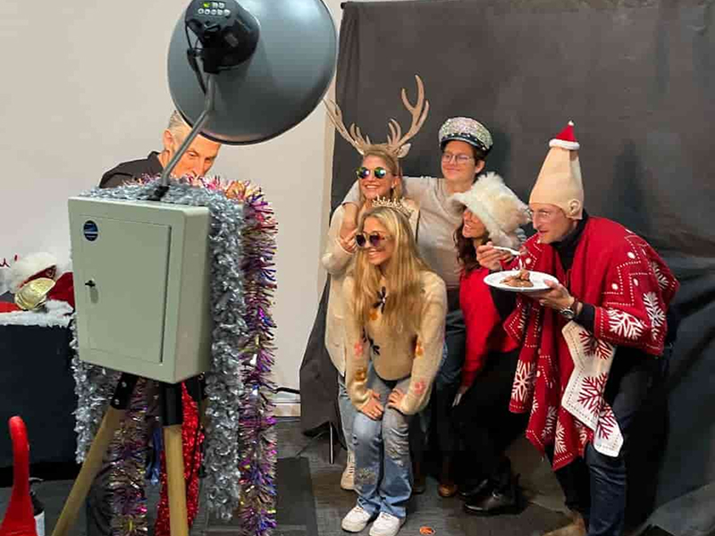 At the digital foundry holiday party, a group of software consultants and family members pose in funny holiday themed hats in front of a photobooth background