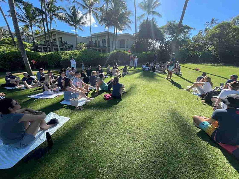 About forty digital foundry software engineers, software consulting staff, other staff and their families sit on a lawn in Hawaii listening to one person speak