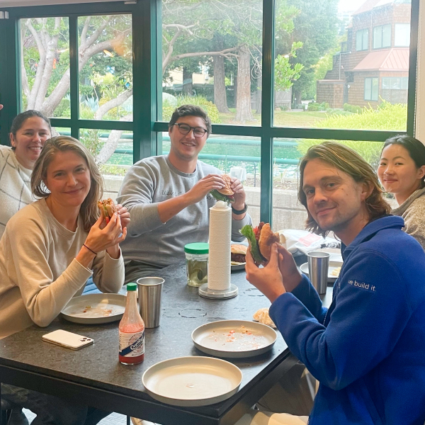 Five Digitial Foundry employees, two men and three women, sit at a lunch table next to a window with plates, cups and hot sauce. They are all eating fried chicken sandwiches from the Sandwich Club.